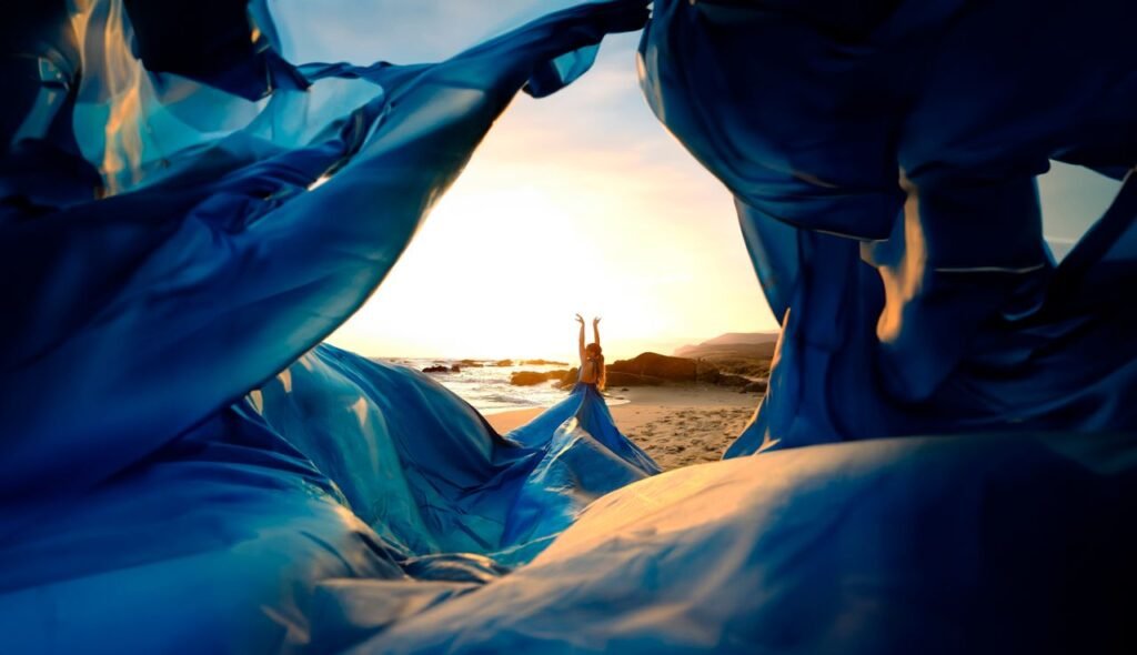 A woman poses elegantly in a blue dress on a beach at sunset, framed by flowing fabric.
